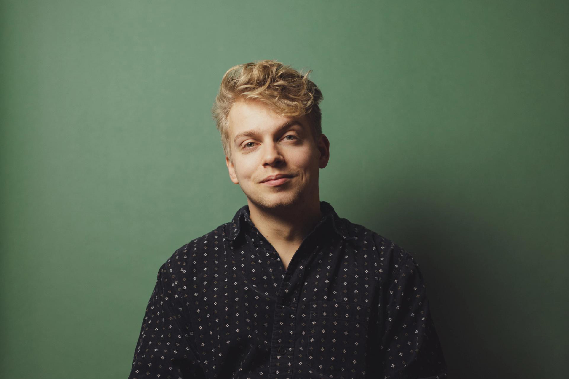 A man with fair hair, wearing a dotted black shirt, smiling.